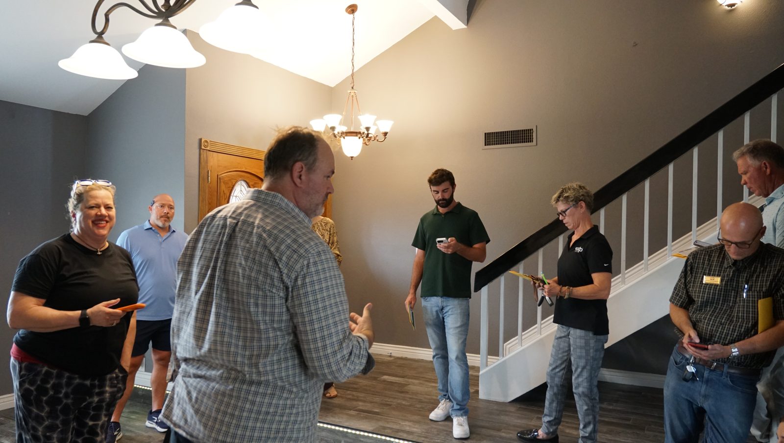 Seven agents gathered in the entry of a home on tour, one mid-sentence, feedback cards in hand.